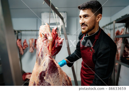 Male butcher standing by meat hanging on hook in meat shop Male butcher standing by meat hanging on hook in meat shop 86633659