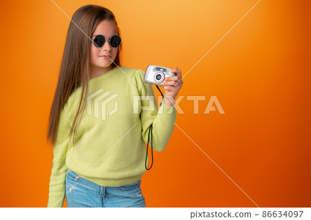 Beautiful teen girl with old photo camera against orange background Beautiful teen girl with old photo camera against orange background 86634097