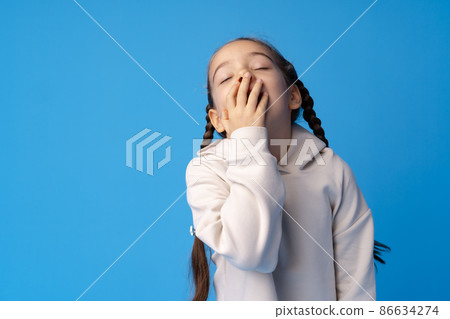 A tired little girl yawning on blue background. A tired little girl yawning on blue background. 86634274