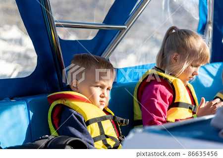Portrait of sad little blond unhappy disappointed caucasian boy wear lifevest feel sick nausea sailing on motor boat sea against sun backlit. Summer water travel with kid vacation recreation 86635456