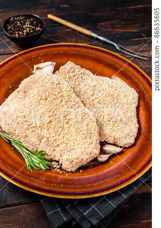 Raw Breaded German Wiener schnitzel on a rustic plate with herbs. Dark wooden background. Top view Raw Breaded German Wiener schnitzel on a rustic plate with herbs. Dark wooden background. Top view 86635605
