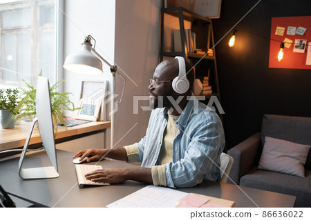 African man in wireless headphones sitting at his workplace in front of the computer monitor and working African man in wireless headphones sitting at his workplace in front of the computer monitor and working 86636022
