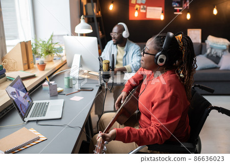 African woman with disability in headphones playing guitar while singing to the microphone during live broadcasting with music director in the background African woman with disability in headphones playing guitar while singing to the microphone during live broadcasting with music director in the background 86636023