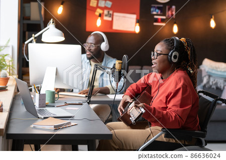 African woman with disability sitting on wheelchair playing guitar and singing a song to the microphone with man using computer in the background African woman with disability sitting on wheelchair playing guitar and singing a song to the microphone with man using computer in the background 86636024