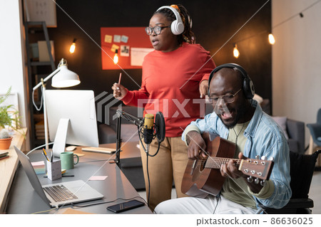 African musician sitting in front of the microphone playing guitar and singing a song together with woman during online broadcasting 86636025