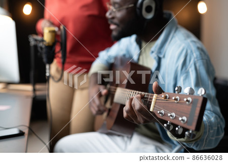 Close-up of African man in headphones playing guitar while sitting opposite the microphone with woman standing in the background Close-up of African man in headphones playing guitar while sitting opposite the microphone with woman standing in the background 86636028