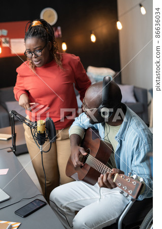 African young woman singing song while her friend playing guitar sitting at the table in front of the microphone during online broadcasting African young woman singing song while her friend playing guitar sitting at the table in front of the microphone during online broadcasting 86636034