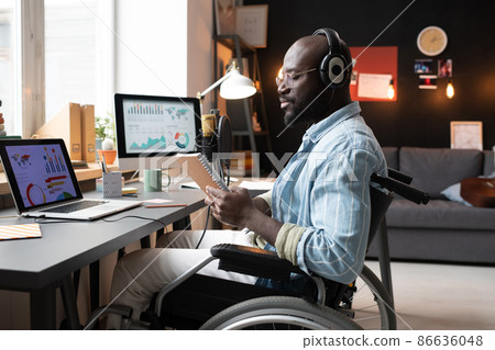 African young man in headphones sitting on wheelchair and reading something in his notebook during his online study at the table 86636048