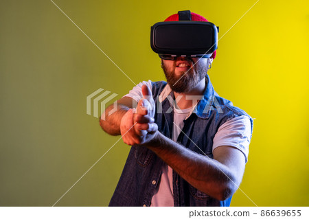 Man with virtual reality glasses on head playing shooter game, pointing finger gun and shooting right on target, wearing denim vest. Indoor studio shot isolated on colorful neon light background. Man with virtual reality glasses on head playing shooter game, pointing finger gun and shooting right on target, wearing denim vest. Indoor studio shot isolated on colorful neon light background. 86639655