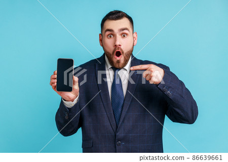 Surprised bearded man wearing official style suit pointing finger at smartphone with white empty display, shocked with new phone features. Indoor studio shot isolated on blue background. Surprised bearded man wearing official style suit pointing finger at smartphone with white empty display, shocked with new phone features. Indoor studio shot isolated on blue background. 86639661