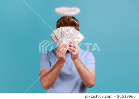 Portrait of unknown anonymous rich angelic man with holy nimbus over head hiding his face behind bunch of dollar banknotes, finance and banking. Indoor studio shot isolated on blue background. Portrait of unknown anonymous rich angelic man with holy nimbus over head hiding his face behind bunch of dollar banknotes, finance and banking. Indoor studio shot isolated on blue background. 86639666