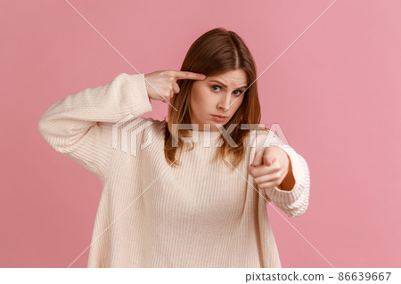Hey, you are kidding. Portrait of blond woman holding finger near head and pointing finger at camera, stupid gesture, wearing white sweater. Indoor studio shot isolated on pink background. 86639667