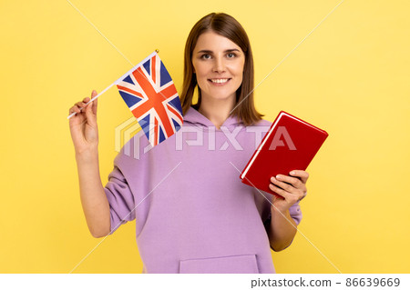 Portrait of attractive student dark haired woman holding book and Great Britain flag, education abroad, wearing purple hoodie. Indoor studio shot isolated on yellow background. Portrait of attractive student dark haired woman holding book and Great Britain flag, education abroad, wearing purple hoodie. Indoor studio shot isolated on yellow background. 86639669
