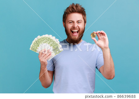 Portrait of positive optimnistic handsome bearded man showing euro banknotes and bitcoin, digital money, electronic commerce, expressing excitement. Indoor studio shot isolated on blue background. Portrait of positive optimnistic handsome bearded man showing euro banknotes and bitcoin, digital money, electronic commerce, expressing excitement. Indoor studio shot isolated on blue background. 86639688