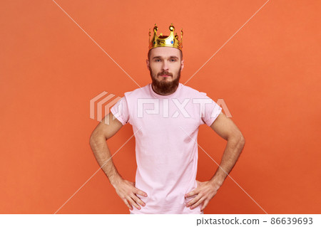 Portrait of bearded man egoistically looking at camera, posing with crown on head, pretending to be king, privileged status, wearing pink T-shirt. Indoor studio shot isolated on orange background. Portrait of bearded man egoistically looking at camera, posing with crown on head, pretending to be king, privileged status, wearing pink T-shirt. Indoor studio shot isolated on orange background. 86639693