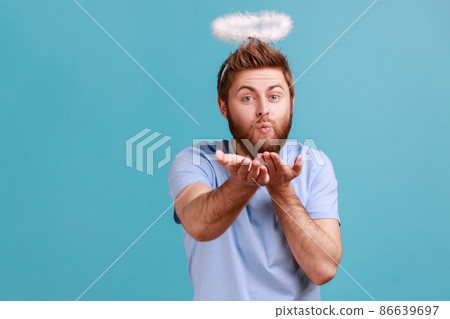 Portrait of handsome romantic young adult bearded angelic man with holy nimbus sending air kiss to camera, flirty angel, valentines day. Indoor studio shot isolated on blue background. 86639697
