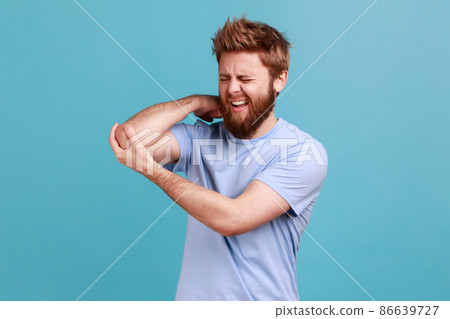 Portrait of bearded man massaging aching elbow, frowning from acute pain, strain and strain problem, trapped nerves, rheumatoid arthritis. Indoor studio shot isolated on blue background. 86639727