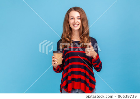 woman wearing striped casual style sweater, holding take away coffee and showing thumb up, recommend coffee house, excellent service.Indoor studio shot isolated on blue background. woman wearing striped casual style sweater, holding take away coffee and showing thumb up, recommend coffee house, excellent service.Indoor studio shot isolated on blue background. 86639739