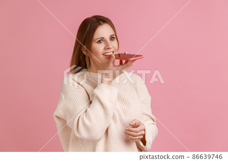 Portrait of optimistic blond woman holding mobile phone in hands, recording voice message or command to assistant, wearing white sweater. Indoor studio shot isolated on pink background. 86639746