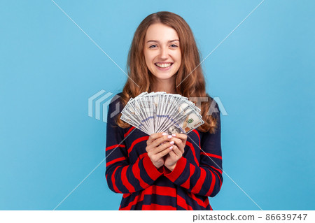 Satisfied woman wearing striped casual style sweater, holding fan of dollar bills and looking ar camera with toothy smile, lottery winner. Indoor studio shot isolated on blue background. 86639747