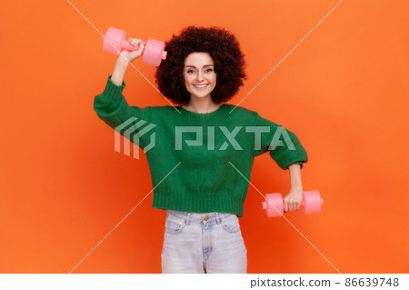 Happy positive sporty woman with Afro hairstyle wearing green casual style sweater doing exercises for biceps, using dumbbells, health care. Indoor studio shot isolated on orange background. 86639748
