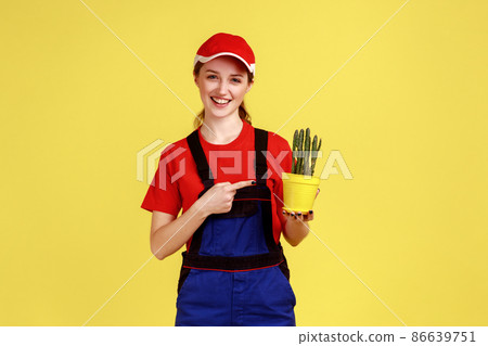 Portrait of happy woman gardener standing pointing at yellow flower pot in hands, showing prickly cactus, wearing overalls and red cap. Indoor studio shot isolated on yellow background. Portrait of happy woman gardener standing pointing at yellow flower pot in hands, showing prickly cactus, wearing overalls and red cap. Indoor studio shot isolated on yellow background. 86639751