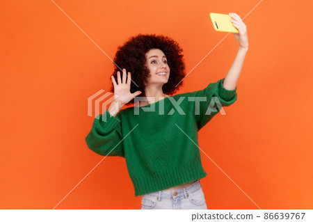 Adorable blogger woman with Afro hairstyle wearing green casual style sweater waving hand to followers while having livestream, greeting new people. Indoor studio shot isolated on orange background. Adorable blogger woman with Afro hairstyle wearing green casual style sweater waving hand to followers while having livestream, greeting new people. Indoor studio shot isolated on orange background. 86639767