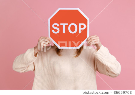 Portrait of unrecognizable woman hiding her face with red sign, campaign against domestic violence, protection of women's rights, wearing white sweater. Indoor studio shot isolated on pink background. Portrait of unrecognizable woman hiding her face with red sign, campaign against domestic violence, protection of women's rights, wearing white sweater. Indoor studio shot isolated on pink background. 86639768