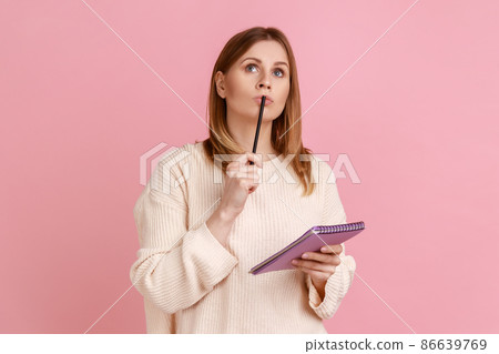 Portrait of pensive blond woman holding pen and paper notebook, being deep in thoughts, writing a composition, wearing white sweater. Indoor studio shot isolated on pink background. Portrait of pensive blond woman holding pen and paper notebook, being deep in thoughts, writing a composition, wearing white sweater. Indoor studio shot isolated on pink background. 86639769
