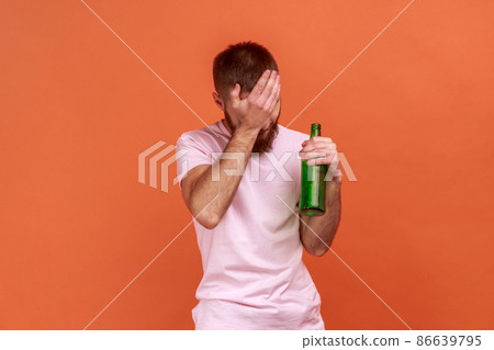 Portrait of upset sad bearded man standing with beer bottle, being drunk, standing covering his face with palm, wearing pink T-shirt. Indoor studio shot isolated on orange background. 86639795