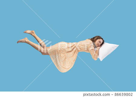 Relaxed girl in yellow dress levitating in mid-air, sleeping on stomach lying comfortable cozy on pillow, keeping eyes closed, watching peaceful dream. indoor studio shot isolated on blue 86639802