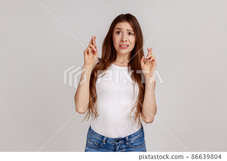 Hopes and wishes for fortune. Portrait of worried woman holding fingers crossed for good luck, expressing great desire to win, wearing white T-shirt. Indoor studio shot isolated on gray background. Hopes and wishes for fortune. Portrait of worried woman holding fingers crossed for good luck, expressing great desire to win, wearing white T-shirt. Indoor studio shot isolated on gray background. 86639804