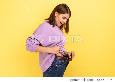 Side view of dark haired happy woman showing successful weight loss, expressing happiness, diet concept, wearing purple hoodie. Indoor studio shot isolated on yellow background. Side view of dark haired happy woman showing successful weight loss, expressing happiness, diet concept, wearing purple hoodie. Indoor studio shot isolated on yellow background. 86639826