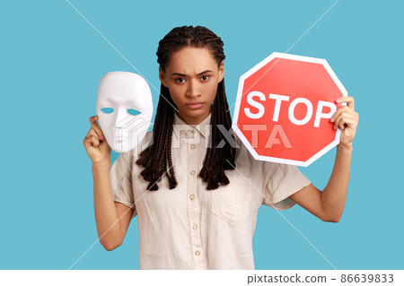 Serious woman with black dreadlocks holding white mask and red stop sign, looking at camera, do not change personality, wearing white shirt. Indoor studio shot isolated on blue background. 86639833