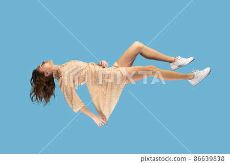 Floating in air. Relaxed girl in yellow dress levitating keeping eyes closed, sleeping while flying mid-air, having comfortable peaceful dream. full length studio shot isolated on blue background 86639838