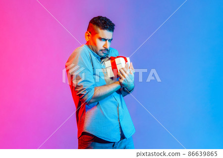 Portrait of man in shirt holding present box in hands, being greedy to share gifts, looking at camera with frowning face. Indoor studio shot isolated on colorful neon light background. 86639865