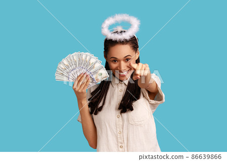 Positive angelic woman with black dreadlocks and halo over head holding fan of dollar bills and pointing finger to camera, choosing lottery winner. Indoor studio shot isolated on blue background. 86639866