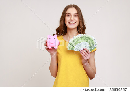 Positive attractive teenager girl with brown hair in yellow casual style holding euro banknotes and piggy bank, savings, high rate deposit. Indoor studio shot isolated on gray background. 86639867