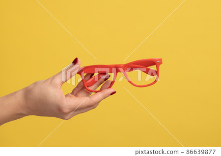 Profile side view closeup of woman hand holding and showing red eyeglasses frame. Indoor studio shot isolated on yellow background. 86639877