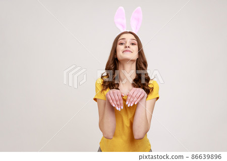 Portrait of joyful beautiful woman of young age in yellow T-shirt behaving childish humorous with bunny ears and hands, entertaining and fooling. Indoor studio shot isolated on gray background. Portrait of joyful beautiful woman of young age in yellow T-shirt behaving childish humorous with bunny ears and hands, entertaining and fooling. Indoor studio shot isolated on gray background. 86639896