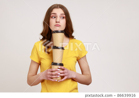 Bored sleepy teenage girl in yellow casual T-shirt holding three coffee, has exhausted expression, bringing drinks in disposable cups. Indoor studio shot isolated on gray background. Bored sleepy teenage girl in yellow casual T-shirt holding three coffee, has exhausted expression, bringing drinks in disposable cups. Indoor studio shot isolated on gray background. 86639916