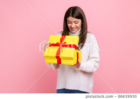 Positive happy dark haired girl looking inside wrapped present box, open gift, expressing positive, wearing white casual style sweater. Indoor studio shot isolated on pink background. 86639930