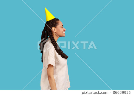 Side view of optimistic pretty woman in party cone hat, looking at camera with toothy smile, rejoicing holiday celebration, wearing white shirt. Indoor studio shot isolated on blue background. 86639935
