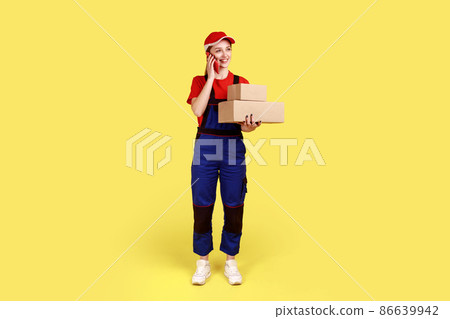 Full length portrait of delivery woman standing with two parcels in hands, talking mobile phone with client, wearing overalls and red cap. Indoor studio shot isolated on yellow background. 86639942
