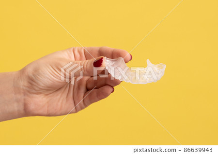 Closeup profile side view portrait of woman hand holding dental aligner retainer at dental clinic for beautiful teeth treatment course. Indoor studio shot isolated on yellow background. 86639943
