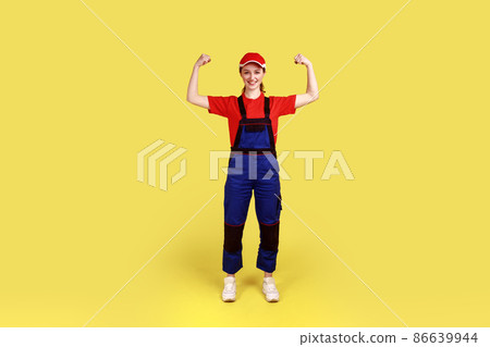 Full length portrait of optimistic woman worker standing and looking at camera with raised arms, showing her power, wearing overalls and red cap. Indoor studio shot isolated on yellow background. Full length portrait of optimistic woman worker standing and looking at camera with raised arms, showing her power, wearing overalls and red cap. Indoor studio shot isolated on yellow background. 86639944