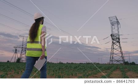 high energized towers against backdrop of sunset in the sky. woman architect hard hat with tablet designing place to control equipment. young woman energy voltage specialist. electric towers at sunset high energized towers against backdrop of sunset in the sky. woman architect hard hat with tablet designing place to control equipment. young woman energy voltage specialist. electric towers at sunset 86641703