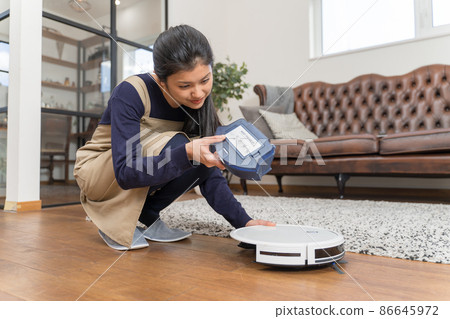 A young woman removing the dust box from a robot vacuum and dumping it A young woman removing the dust box from a robot vacuum and dumping it 86645972