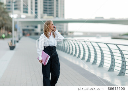 A blonde businesswoman holds a folder and talks on her smartphone in the city 86652600