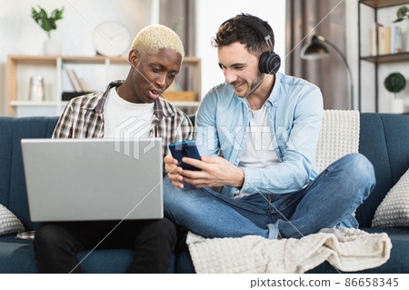 Multiracial same sex couple resting on couch with gadgets Multiracial same sex couple resting on couch with gadgets 86658345